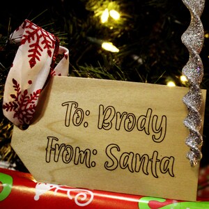 May include: A wooden gift tag with the text "To: Brody From: Santa" written in black cursive. The tag is decorated with a red and white snowflake ribbon.