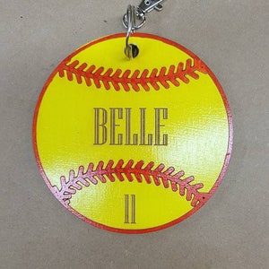 May include: Yellow softball keychain with red stitching and the name "BELLE" and the number "II" written in gold. The keychain has a silver metal clasp.
