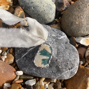 以下が含まれることがあります： ターコイズとクリーム色の模様が入ったシーグラスのペンダントネックレス。シルバーカラーのメタルフレームにセットされています。白いリボンから吊り下げられ、小石に囲まれた灰色の石の上に置かれています。