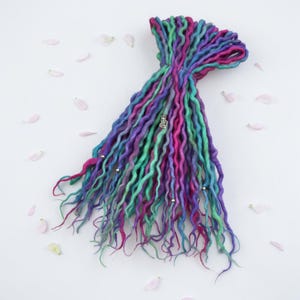 May include: A bundle of colorful dreadlocks in shades of purple, teal, and pink, tied together with a thin brown string. The dreadlocks are adorned with small silver beads and a decorative charm. Scattered around the dreadlocks are pink flower petals.