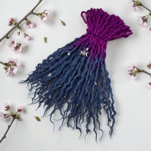 Peut inclure: Une mèche de dreadlocks faits à la main, dégradé de violet à bleu marine. Les dreads sont ornées de breloques argentées, dont des étoiles et des croissants de lune. En arrière-plan, des branches de fleurs de cerisier.