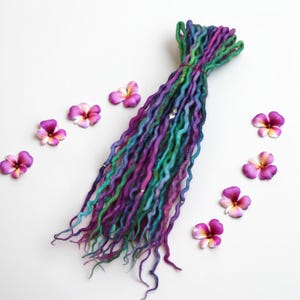 Può includere: Un fascio di dreadlocks colorati in tonalità di verde, blu e viola, adornati con piccoli accenti argentati. I dreadlocks sono disposti su una superficie bianca, circondati da diversi fiori rosa e viola.