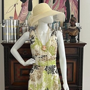 May include: A sleeveless floor-length dress featuring a cream base with a floral pattern in shades of lime green, brown, and black. The dress has a V-neckline, a gathered waist, and is accessorized with a long white pearl necklace and a cameo pendant.
