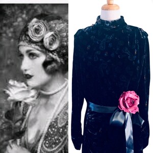 May include: A dark navy blue long-sleeved dress featuring a subtle tonal paisley pattern throughout the velvet fabric. The dress has a high neckline and is accented at the waist with a dark satin ribbon sash and a large, vibrant pink fabric rose.