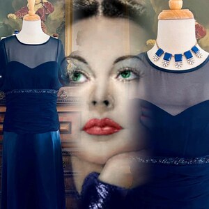 May include: Navy blue evening gown with a sheer overlay on the bodice and sleeves. The dress features a sweetheart neckline and a beaded waist detail. A matching blue necklace with square stones is displayed on a mannequin.