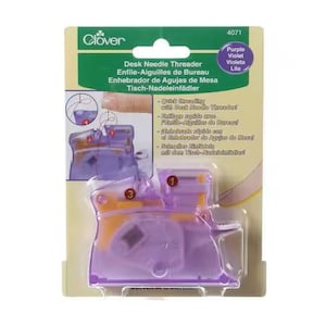 May include: A purple and yellow desk needle threader in its original packaging. The packaging is green and white with the Clover logo and product information. The text on the packaging includes "Desk Needle Threader" and "Purple Violet Lila".