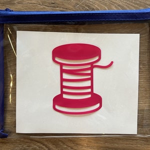 Könnte beinhalten: Eine durchsichtige, mit einem Reißverschluss verschlossene Plastiktasche mit einer weißen Karte im Inneren. Die Karte zeigt eine rosa Silhouette einer Garnrolle.