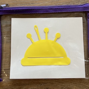 Könnte beinhalten: Ein gelber Nadelkissen mit einer Nadel und vier Stecknadeln, in einer durchsichtigen Plastiktasche mit einem violetten Reißverschluss.