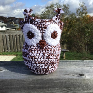 May include: A brown and white crocheted owl with large white eyes and a brown beak. The owl is sitting on a wooden surface.