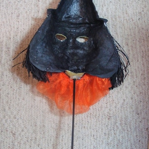 May include: A handmade Halloween mask featuring a black witch's hat and face with eye holes. The mask has an orange tulle collar and is attached to a black stick. The mask is likely used for costume or decorative purposes.