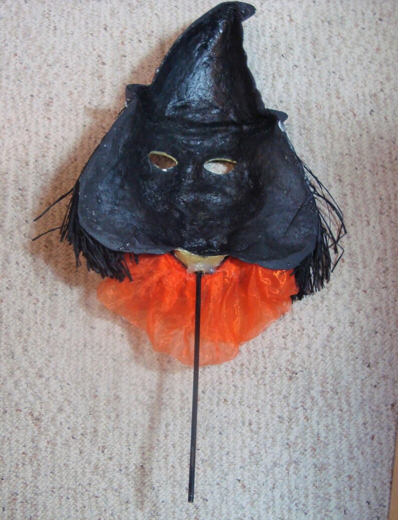 May include: A handmade Halloween mask featuring a black witch's hat and face with eye holes. The mask has an orange tulle collar and is attached to a black stick. The mask is likely used for costume or decorative purposes.