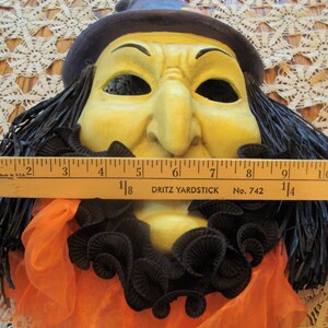May include: A vintage Halloween witch mask with a yellow face, black hair, and a purple hat. The mask has a black ruffled collar and orange fabric. The mask measures approximately 11 inches across.