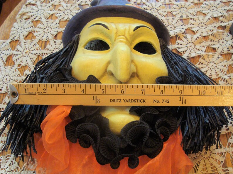 May include: A vintage Halloween witch mask with a yellow face, black hair, and a purple hat. The mask has a black ruffled collar and orange fabric. The mask measures approximately 11 inches across.