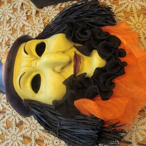 May include: A vintage Halloween mask featuring a witch design. The mask has a yellow face with black hair, a purple hat, and an orange fabric collar. The mask is likely made of plastic or a similar material.