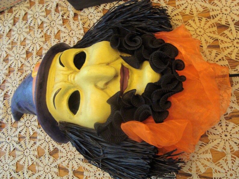 May include: A vintage Halloween mask featuring a witch design. The mask has a yellow face with black hair, a purple hat, and an orange fabric collar. The mask is likely made of plastic or a similar material.
