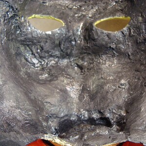 May include: A close-up of a textured, metallic mask with eye holes. The mask is primarily silver with black accents, and the eye holes are lined with yellow. The bottom of the mask has a red accent.