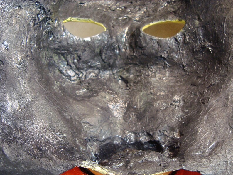 May include: A close-up of a textured, metallic mask with eye holes. The mask is primarily silver with black accents, and the eye holes are lined with yellow. The bottom of the mask has a red accent.