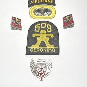 May include: A collection of military insignia including an Airborne tab, a parachute badge on a yellow backing, a 509 Geronimo patch, two shield-shaped pins with the text ALL THE WAY, and a silver-toned winged parachute pin with a red star emblem.