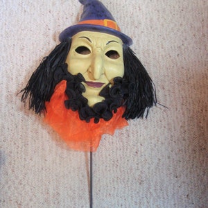 May include: A Halloween witch mask with a pale face, black hair, and a purple hat with an orange band. The mask has a long nose and a black ruffled collar over an orange fabric.