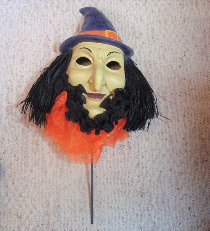 May include: A Halloween witch mask with a pale face, black hair, and a purple hat with an orange band. The mask has a long nose and a black ruffled collar over an orange fabric.