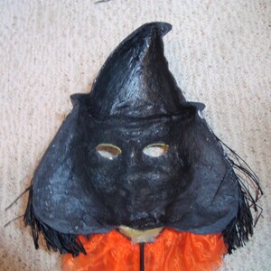 May include: A black witch mask with a pointed hat and eye holes. The mask has a textured surface and is trimmed with black fringe. An orange fabric is visible below the mask, suggesting a costume.