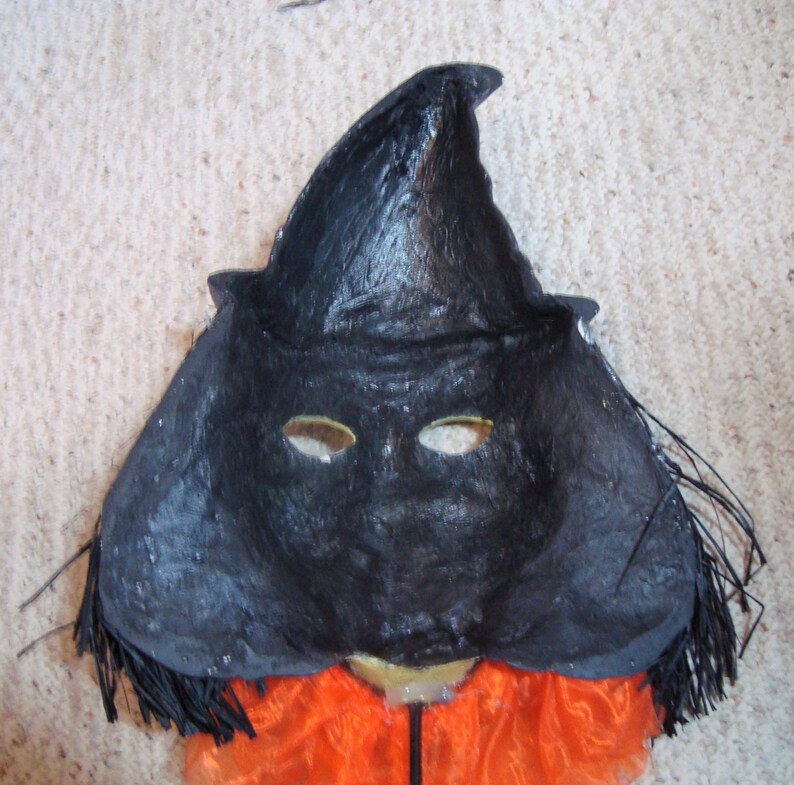 May include: A black witch mask with a pointed hat and eye holes. The mask has a textured surface and is trimmed with black fringe. An orange fabric is visible below the mask, suggesting a costume.