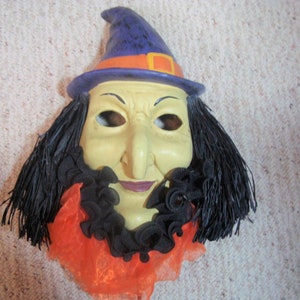 May include: A Halloween witch mask with a yellow face, long black hair, and a purple hat with an orange band. The mask has a long nose, a black ruffled collar, and an orange and black neckpiece.