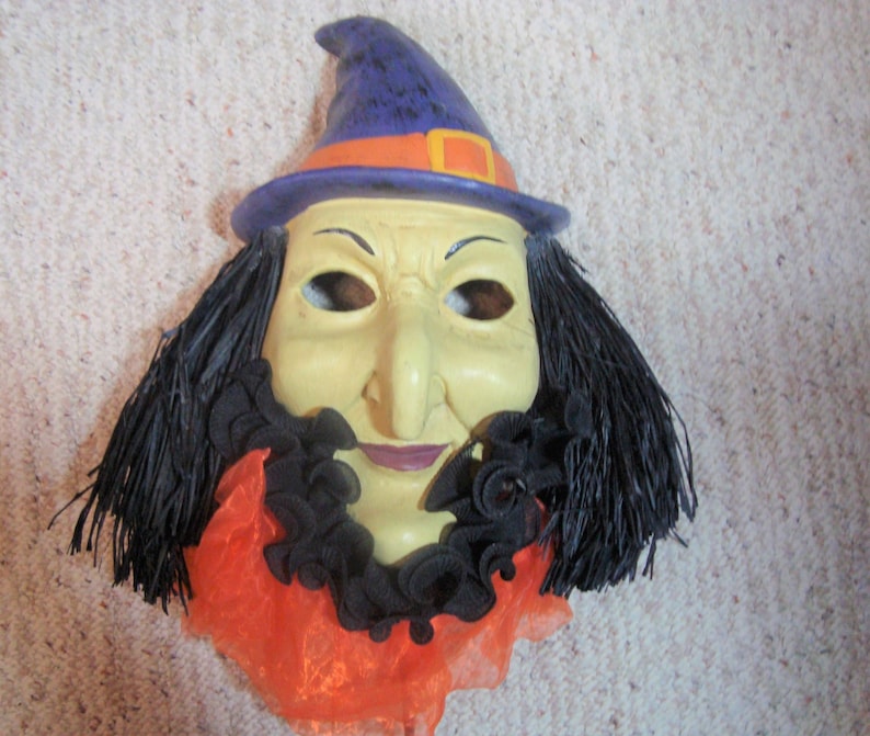 May include: A Halloween witch mask with a yellow face, long black hair, and a purple hat with an orange band. The mask has a long nose, a black ruffled collar, and an orange and black neckpiece.