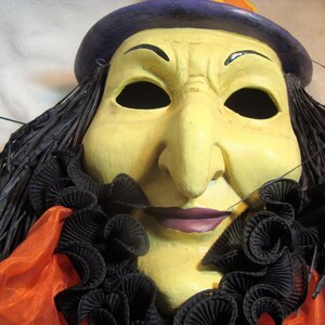 May include: A Halloween witch mask with a yellow face, black hair, and a purple hat. The mask has a large nose, black eyes, and a purple mouth. It is decorated with a black ruffled collar and orange fabric.