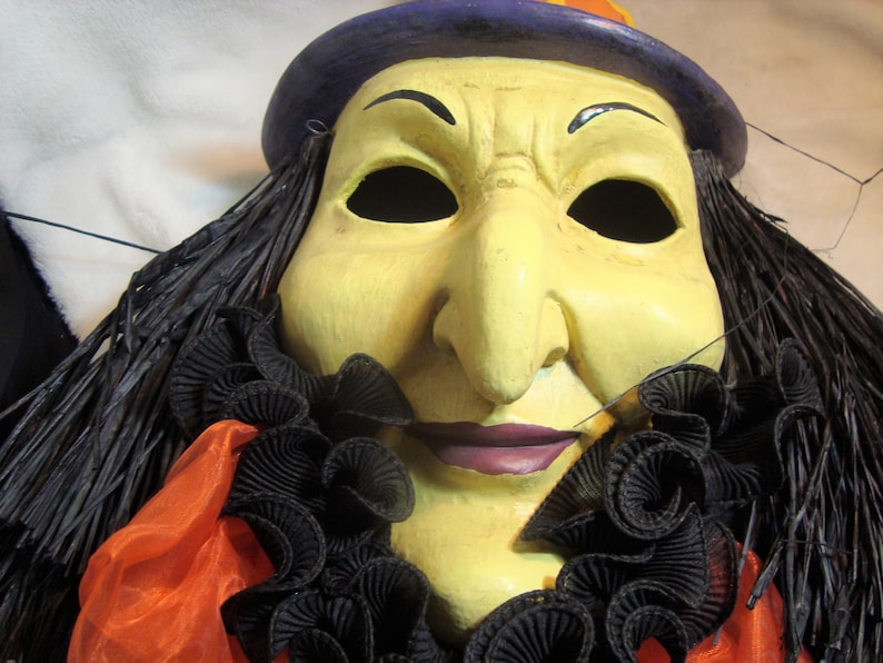 May include: A Halloween witch mask with a yellow face, black hair, and a purple hat. The mask has a large nose, black eyes, and a purple mouth. It is decorated with a black ruffled collar and orange fabric.