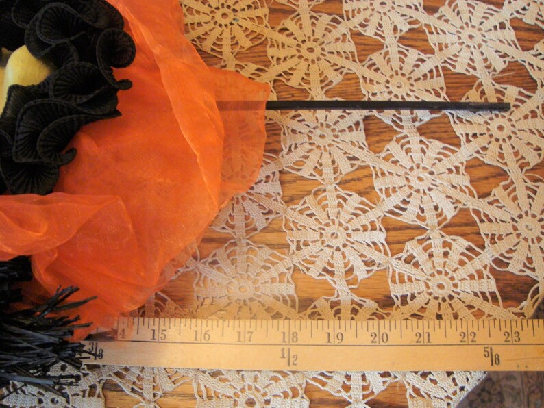 May include: A Halloween-themed image featuring a black and orange witch's broom with a black handle. The broom is adorned with black ribbon and orange fabric. A ruler is placed near the broom, measuring approximately 23 inches.
