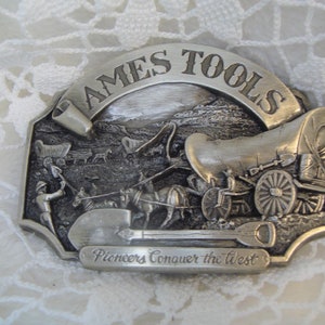 May include: A silver belt buckle with a scene of a covered wagon train and the text "AMES TOOLS Pioneers Conquer the West".