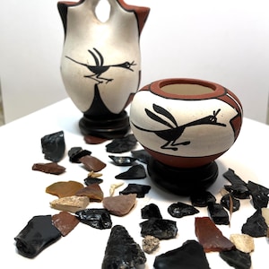 May include: Two small ceramic pots with black and white bird designs. The pots are on a white surface with a scattering of black and brown stones.