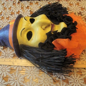 May include: A vintage Halloween mask with a witch-like appearance. The mask features a yellow face with black hair, a purple hat with an orange band, and orange fabric. The mask is lying on a lace tablecloth.