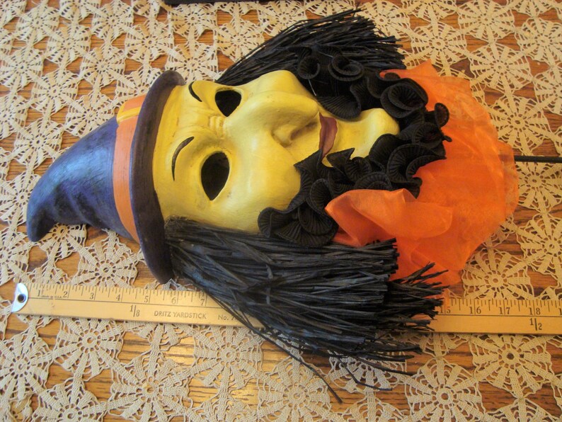 May include: A vintage Halloween mask with a witch-like appearance. The mask features a yellow face with black hair, a purple hat with an orange band, and orange fabric. The mask is lying on a lace tablecloth.