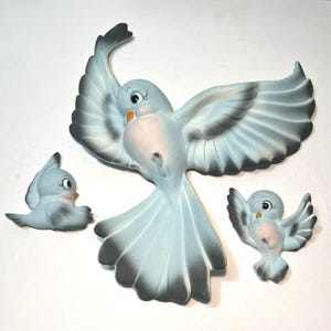 May include: A set of three wall-mounted bird figurines in light blue with white bellies and dark gray wing and tail tips. The largest bird is depicted in flight with wings spread wide, while the two smaller birds are shown in different poses.
