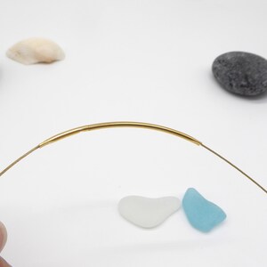 May include: A delicate gold-colored necklace with a curved bar pendant. The necklace is displayed on a white surface with small decorative elements, including sea glass and pebbles. The necklace is a simple, elegant piece of jewelry.