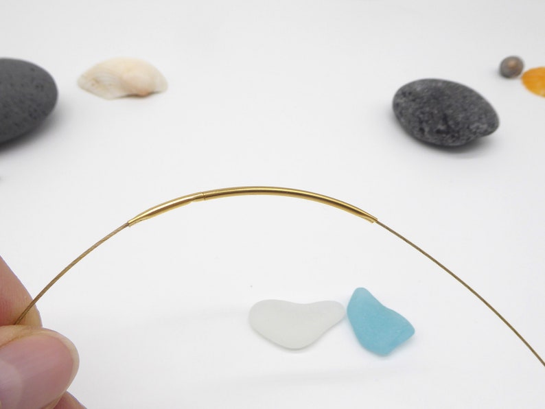 May include: A delicate gold-colored necklace with a curved bar pendant. The necklace is displayed on a white surface with small decorative elements, including sea glass and pebbles. The necklace is a simple, elegant piece of jewelry.