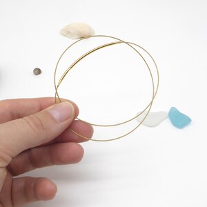 May include: Delicate gold-colored wire bracelets. The bracelets are held in a hand against a white background, with small seashells and sea glass scattered around. The bracelets have a minimalist design.