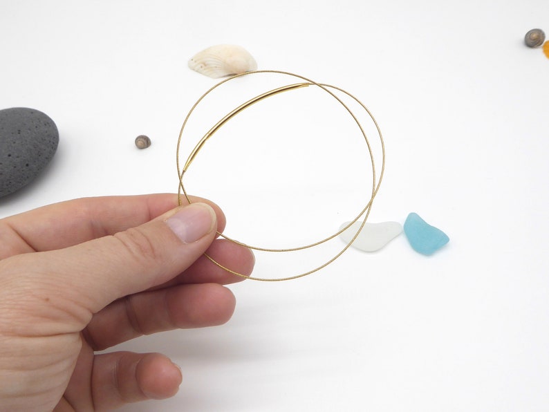May include: Delicate gold-colored wire bracelets. The bracelets are held in a hand against a white background, with small seashells and sea glass scattered around. The bracelets have a minimalist design.
