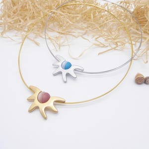 Puede incluir: Dos collares con un colgante de metal en forma de sol sobre un collar de alambre fino. Uno es de tono dorado con un centro circular a rayas rosas y blancas, y el otro es de tono plateado con un centro circular a rayas azules y blancas.
