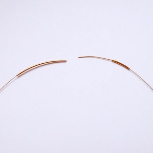 May include: Two pieces of thin, curved, copper-colored wire, each with a slightly thicker section. The wires are positioned against a white background, suggesting a craft or jewelry-making theme. The ends of the wires are cut.