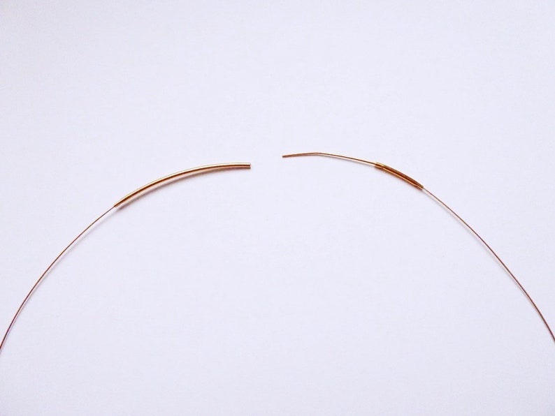 May include: Two pieces of thin, curved, copper-colored wire, each with a slightly thicker section. The wires are positioned against a white background, suggesting a craft or jewelry-making theme. The ends of the wires are cut.