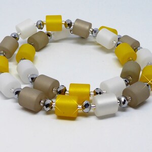 May include: A necklace featuring cylindrical beads in white, yellow, and taupe, interspersed with faceted silver beads. The necklace is arranged in a circular pattern, showcasing the color variations and textures of the beads.