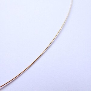 May include: A delicate gold-colored necklace chain against a white background. The chain is thin and appears to be made of metal. The image is a close-up, highlighting the chain's texture and color. The chain is slightly curved, suggesting it is flexible.