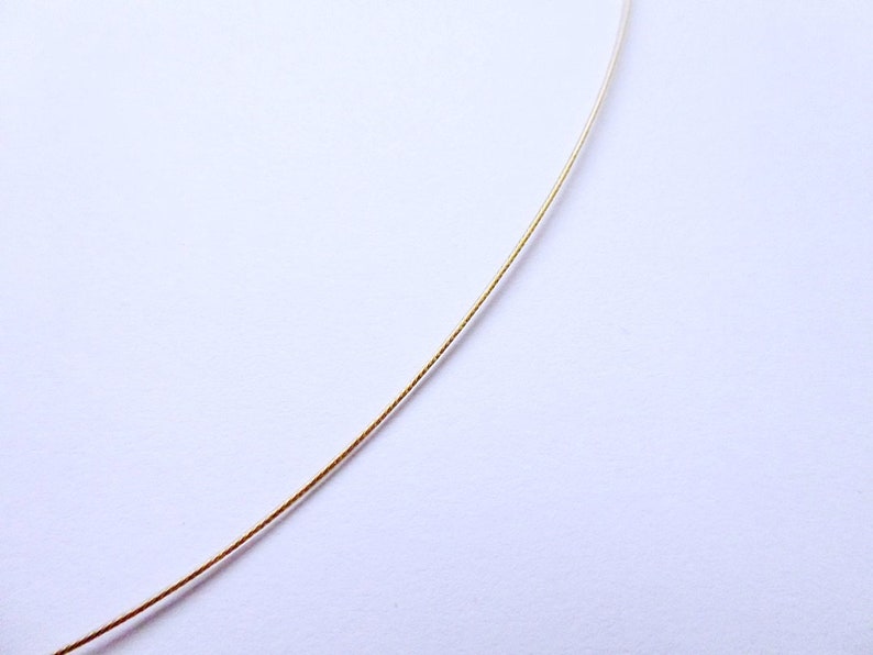 May include: A delicate gold-colored necklace chain against a white background. The chain is thin and appears to be made of metal. The image is a close-up, highlighting the chain's texture and color. The chain is slightly curved, suggesting it is flexible.