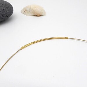 May include: A delicate gold-colored necklace with a thin, flexible wire. The necklace features a curved, tubular gold-toned centerpiece. The design is minimalist and elegant, suitable for everyday wear. The necklace is shown against a white background.