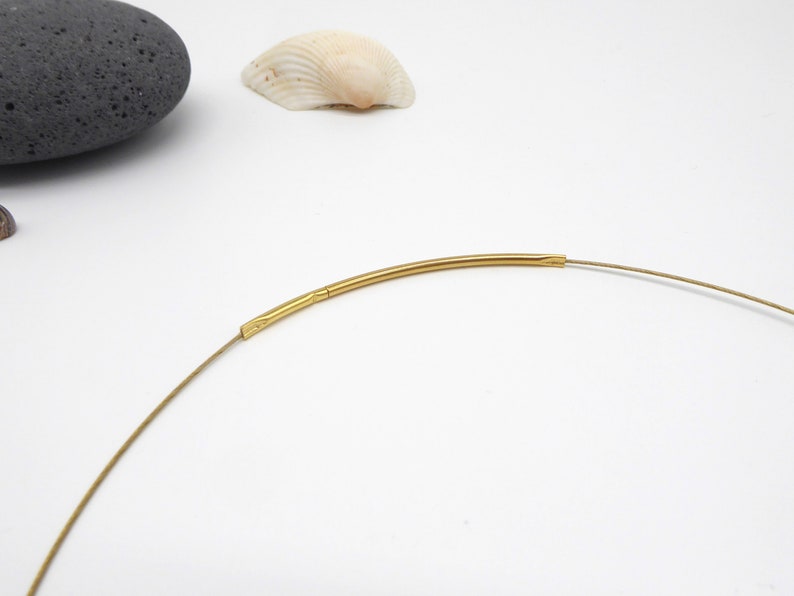 May include: A delicate gold-colored necklace with a thin, flexible wire. The necklace features a curved, tubular gold-toned centerpiece. The design is minimalist and elegant, suitable for everyday wear. The necklace is shown against a white background.