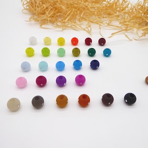 Könnte beinhalten: Eine Sammlung farbenfroher runder Perlen in verschiedenen Farbtönen, darunter Rosa, Blau, Grün und Braun. Jede Perle hat einen kleinen, funkelnden Akzent. Die Perlen sind auf einer weißen Oberfläche angeordnet, mit dekorativen Elementen im Hintergrund.