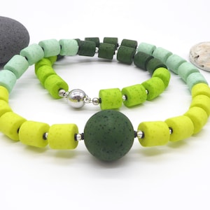 May include: A vibrant necklace featuring a gradient of green hues, from light yellow-green to deep forest green. The necklace is composed of cylindrical beads and a central spherical bead, all connected with silver-toned findings. The necklace is a unique piece of jewelry.